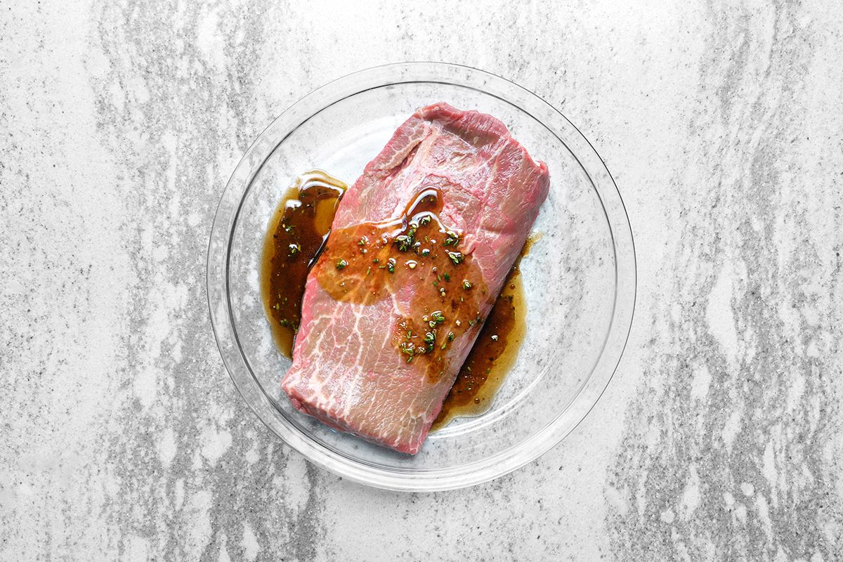 A raw piece of meat marinating in a glass bowl with a dark liquid sauce, sprinkled with herbs. The bowl is placed on a light gray textured surface.