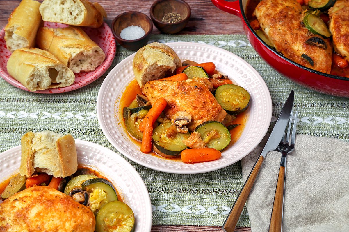 Plate with roasted chicken, zucchini, carrots, and mushrooms, served with slices of baguette. Additional bread and a red pot of food are seen in the background. Salt and pepper are in small bowls nearby, with a fork and knife on a napkin.