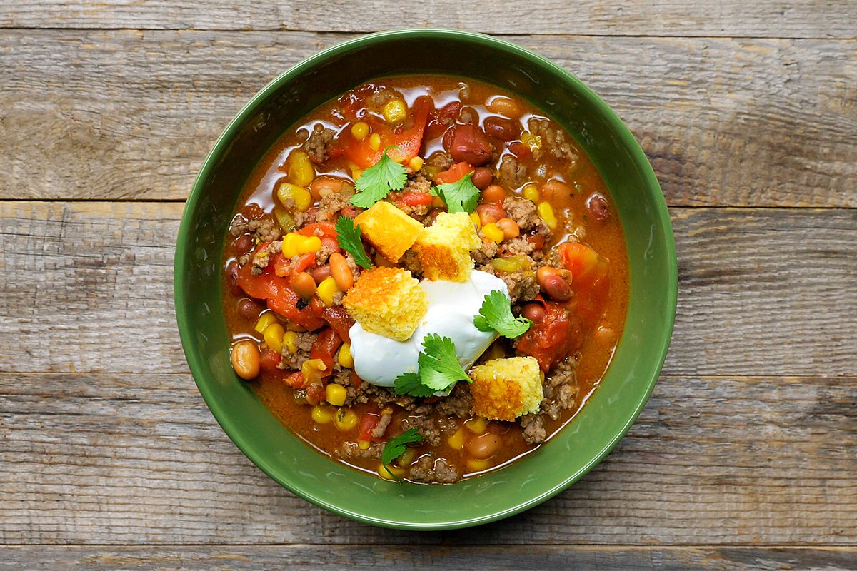 A green bowl filled with chili, featuring ground meat, corn, beans, and diced tomatoes. Topped with a dollop of sour cream, croutons, and garnished with fresh cilantro. Bowl rests on a wooden surface.