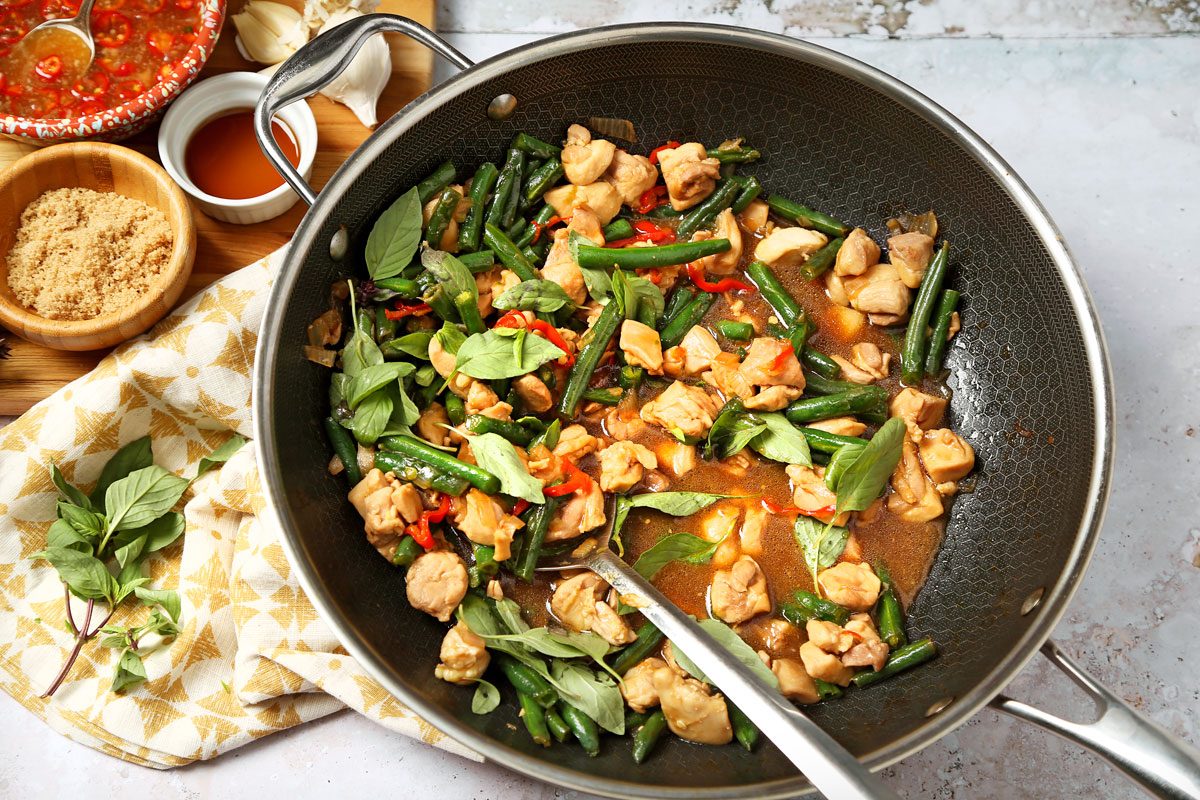 A pan filled with stir-fried chicken, green beans, and red chili peppers, garnished with basil leaves. Next to the pan are bowls of brown sugar, fish sauce, and garlic, placed on a patterned cloth. Thai Basil Chicken