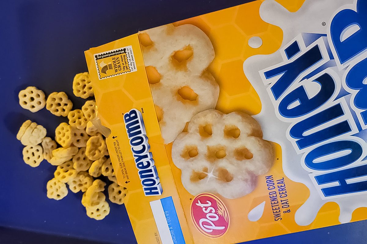 A yellow box of Post Honeycomb cereal is tipped over on a blue surface, with some hexagonal cereal pieces spilling out of the box.