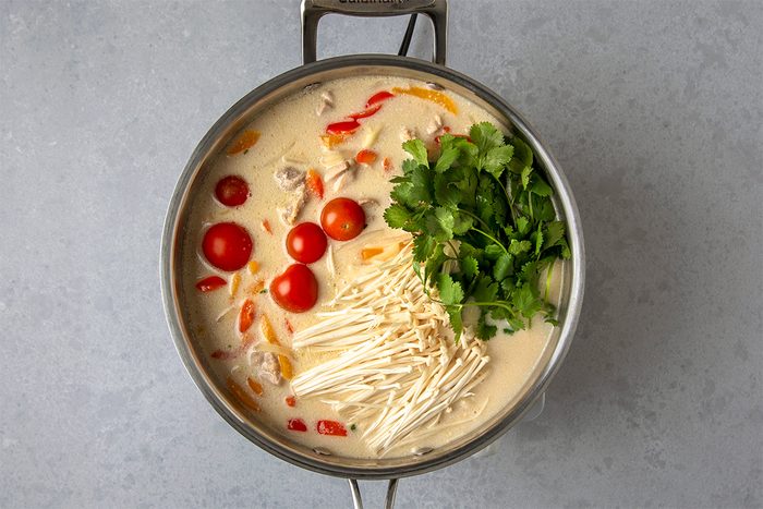 A pot of soup with coconut milk, cherry tomatoes, sliced bell peppers, enoki mushrooms, and fresh cilantro. The soup appears creamy and colorful, with ingredients arranged visibly from above.