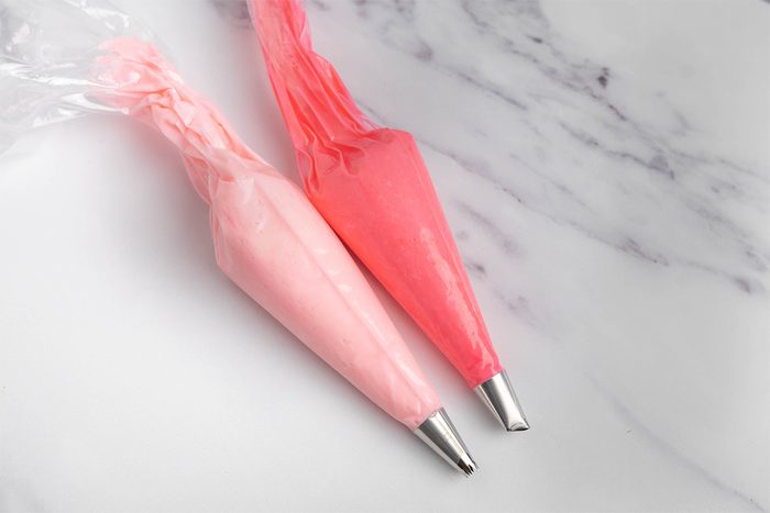 Two piping bags filled with pink and red icing are placed on a white marble surface. The bags are fitted with metal tips, ready for decorating.