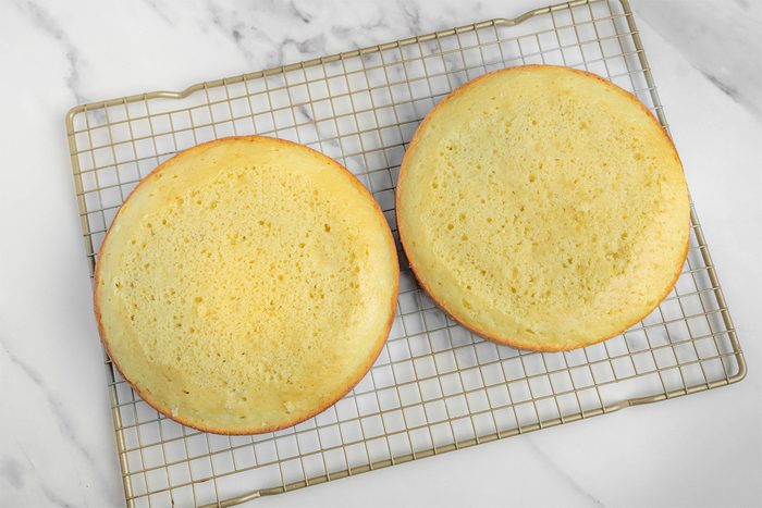 Two round yellow cakes cooling on a wire rack over a marbled countertop. The cakes are evenly baked, with a light golden color.