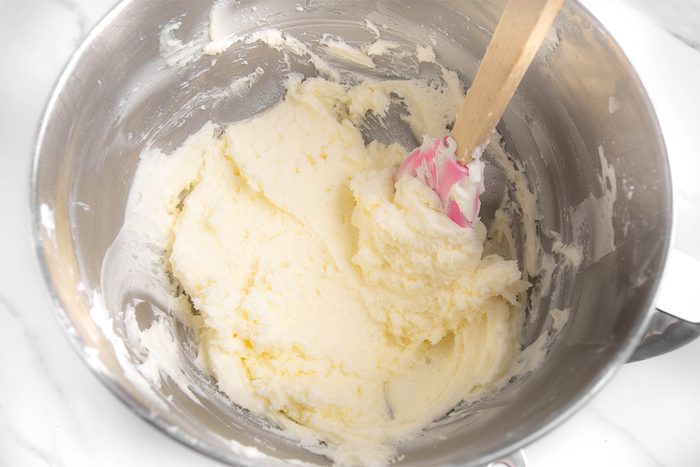 A mixing bowl filled with creamy, pale yellow butter and sugar mixture. A spatula with a pink and white handle rests inside the bowl. The bowl is placed on a white marble surface.