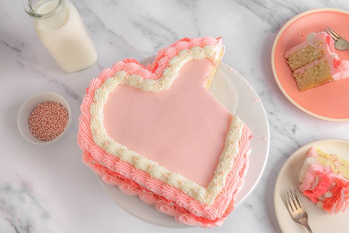 Heart-shaped pink cake with white and pink piped frosting on a white plate. A bottle of milk and a bowl of pink sprinkles sit nearby. A slice of the cake is served on a pink plate with a fork, all placed on a marble surface.