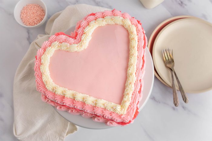 Heart-shaped pink cake with white and darker pink frosting edges on a round cake stand. Beside it, there's a small bowl of pink sprinkles, plates, and forks on a light-colored surface.