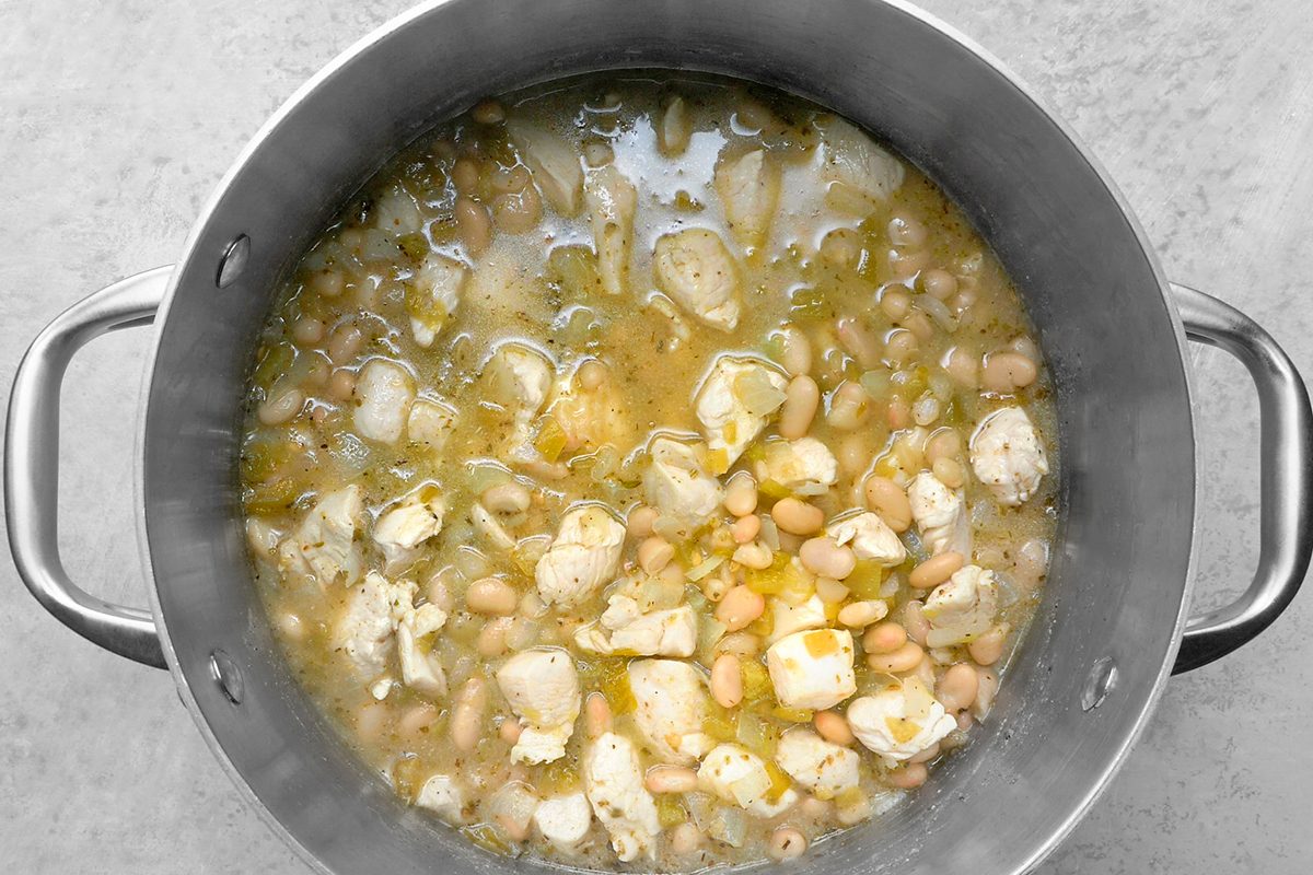 A pot of chicken and white bean soup with visible chunks of chicken, beans, and a light broth. The soup is simmering on a stove with handles on each side of the pot. The background is a light gray countertop.