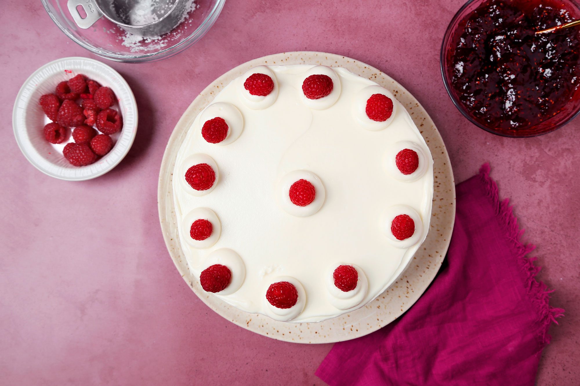 White chocolate Raspberry Cake shot on a magenta surface. Cake assembled on a serving patter Whipped cream and fresh raspberries on the cooled cake.