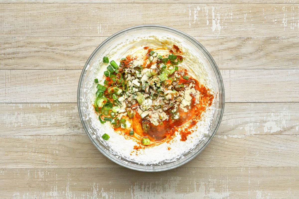 overhead shot of a glass bowl filled with a creamy white mixture, topped with chopped green onions, a sprinkle of red powder, and a few small brown flakes; the bowl sits on a light wood surface