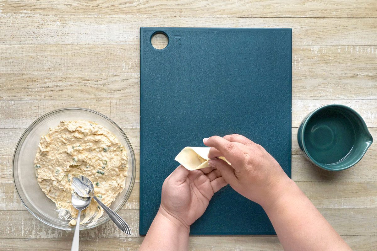 overhead shot of a person preparing food on a wooden table; a glass bowl filled with creamy, white filling with green bits is to the left of the image, and two silver spoons are resting in the bowl; a dark blue cutting board is to the right of the image with a hand preparing a light-colored dough wrapper; a small dark blue bowl filled with water is to the right of the cutting board