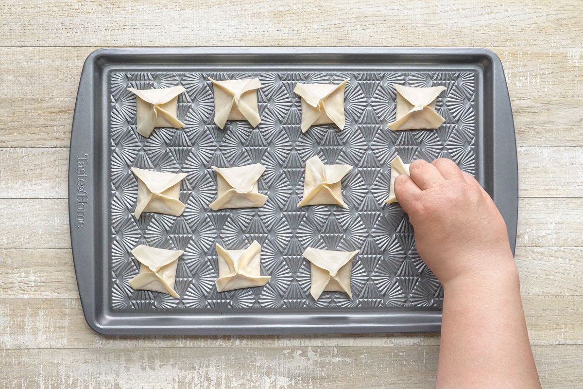 overhead shot of a baking sheet filled with 9 square-shaped dumplings; a hand is placing the last dumpling onto the sheet; the sheet has a gray background with a pattern of radiating lines
