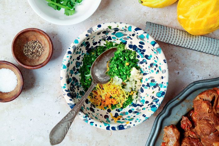 overhead shot of a white countertop with various ingredients and cooking items arranged on it; there is a bowl with green parsley, another bowl with salt, another bowl with pepper, a lemon zester and two lemons, a small bowl with chopped parsley, lemon zest, and garlic, a spoon, and a plate with braised meat