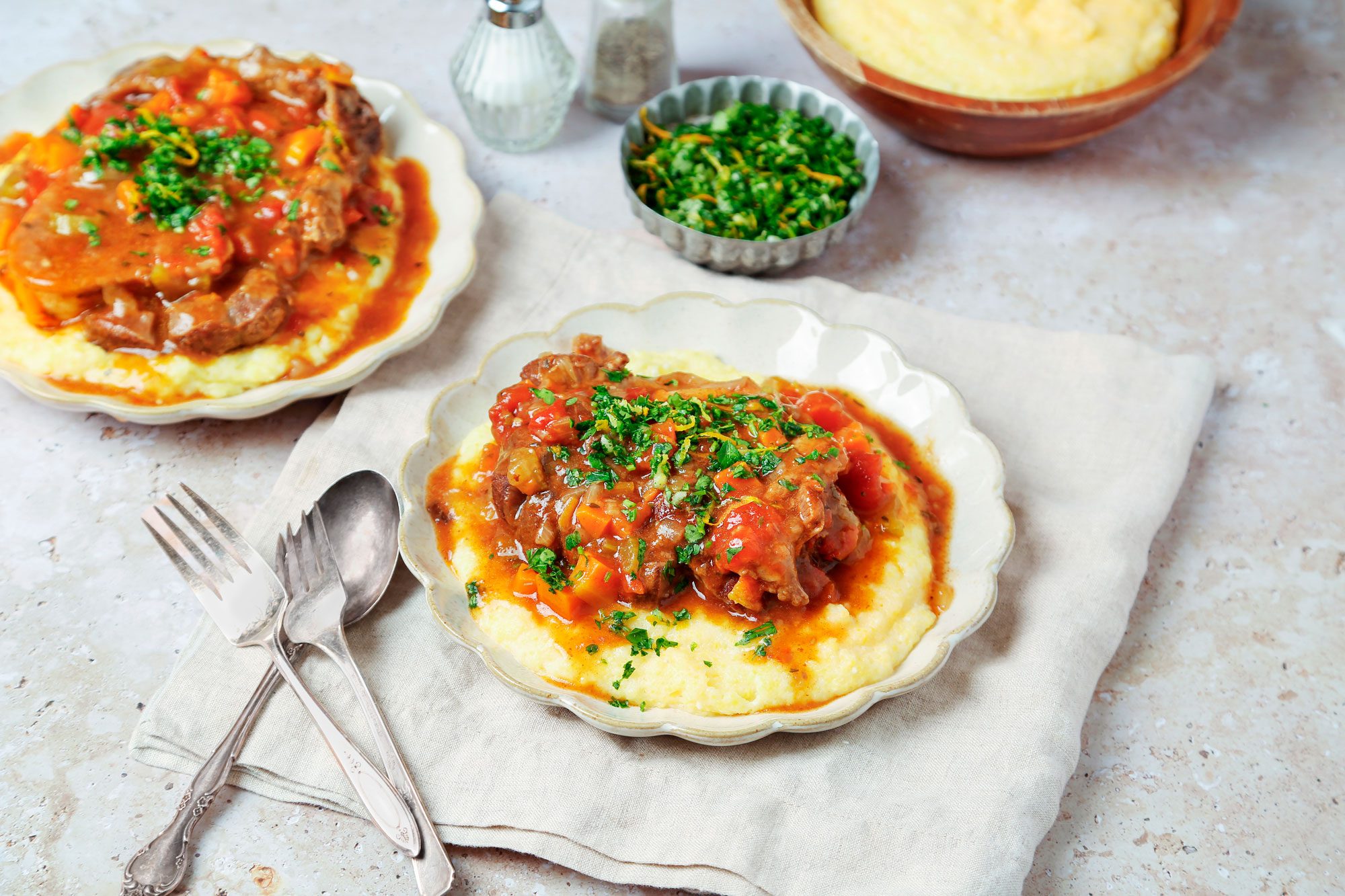 3/4th shot of osso buco with polenta, garnished with parsley and served on a white plate, a napkin, and a light gray marble counter top, two salt shakers are on the counter in the background of the image and a small bowl of chopped herbs, there are also two forks on the napkin