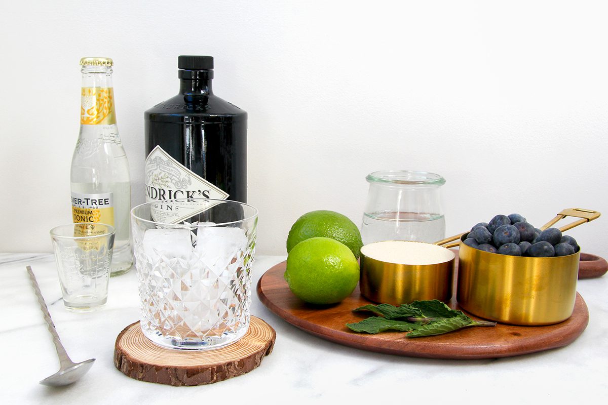 A marble countertop holds ingredients for a cocktail: a bottle of tonic water, gin, a glass with ice on a wooden coaster, two limes, a golden bowl of blueberries, fresh mint, a spoon, and a jar of clear liquid on a wooden board.