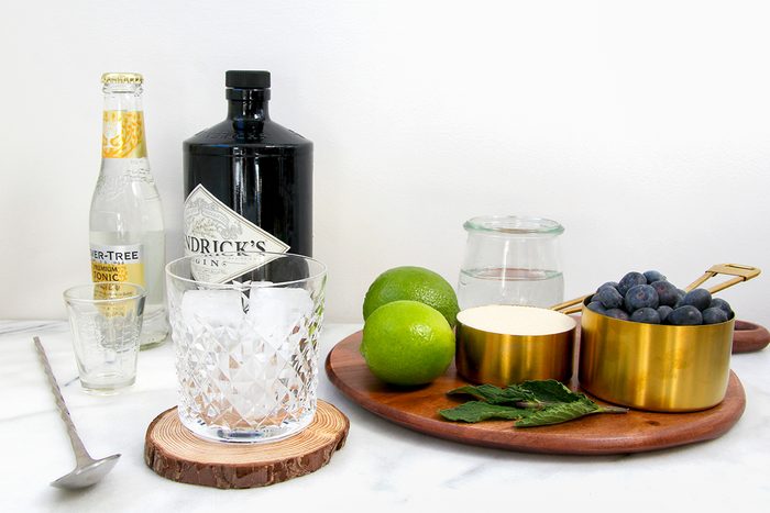A marble countertop holds ingredients for a cocktail: a bottle of tonic water, gin, a glass with ice on a wooden coaster, two limes, a golden bowl of blueberries, fresh mint, a spoon, and a jar of clear liquid on a wooden board.