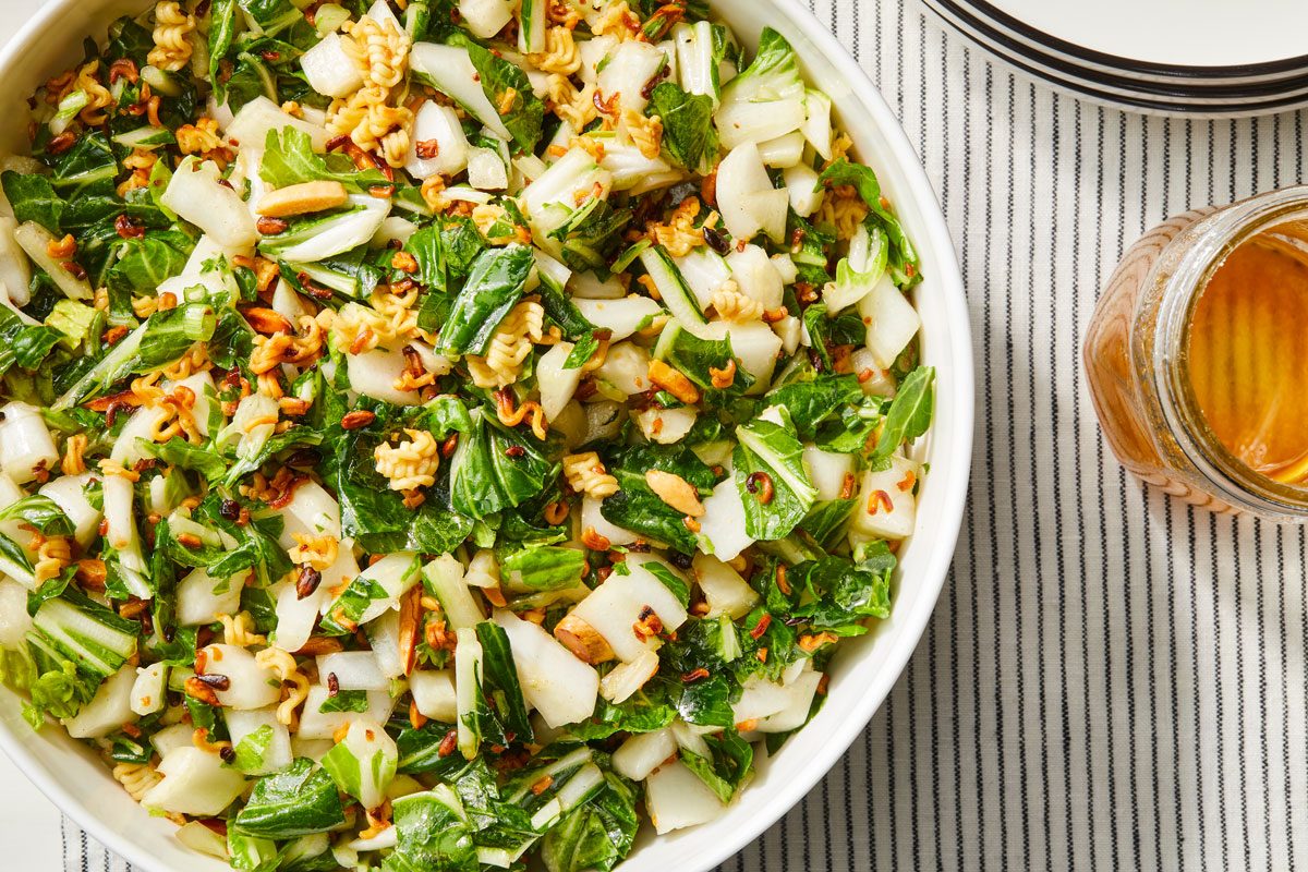 Overhead shot of Bok Choy Salad; in a white bowl; a glass pitcher or jar in the background; and empty plates arranged on a white and grey striped tablecloth;