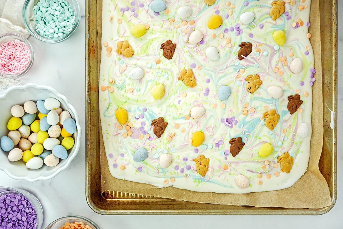 A baking tray with white chocolate bark topped with colorful candy eggs and teddy bear-shaped cookies. Surrounding the tray are small bowls filled with more colorful candies and sprinkles.