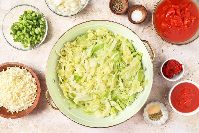 A bowl of shredded cabbage is surrounded by bowls of diced green peppers, chopped onions, diced tomatoes, grated cheese, and seasonings on a light-colored countertop.