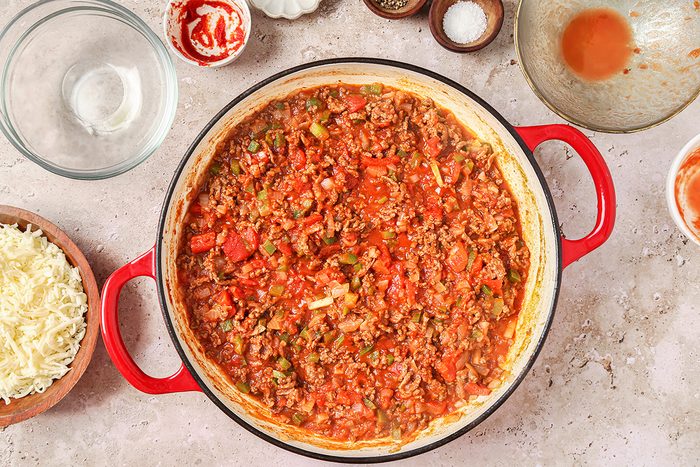 A red pot filled with chunky meat sauce, including visible pieces of tomatoes and green peppers, is placed on a speckled countertop. Surrounding the pot are empty bowls and a dish of shredded cheese.