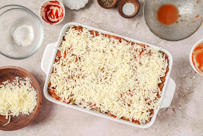 A casserole dish filled with lasagna is topped with grated cheese. Surrounding the dish are empty bowls, a bowl of grated cheese, some tomato sauce, and seasonings on a light countertop.