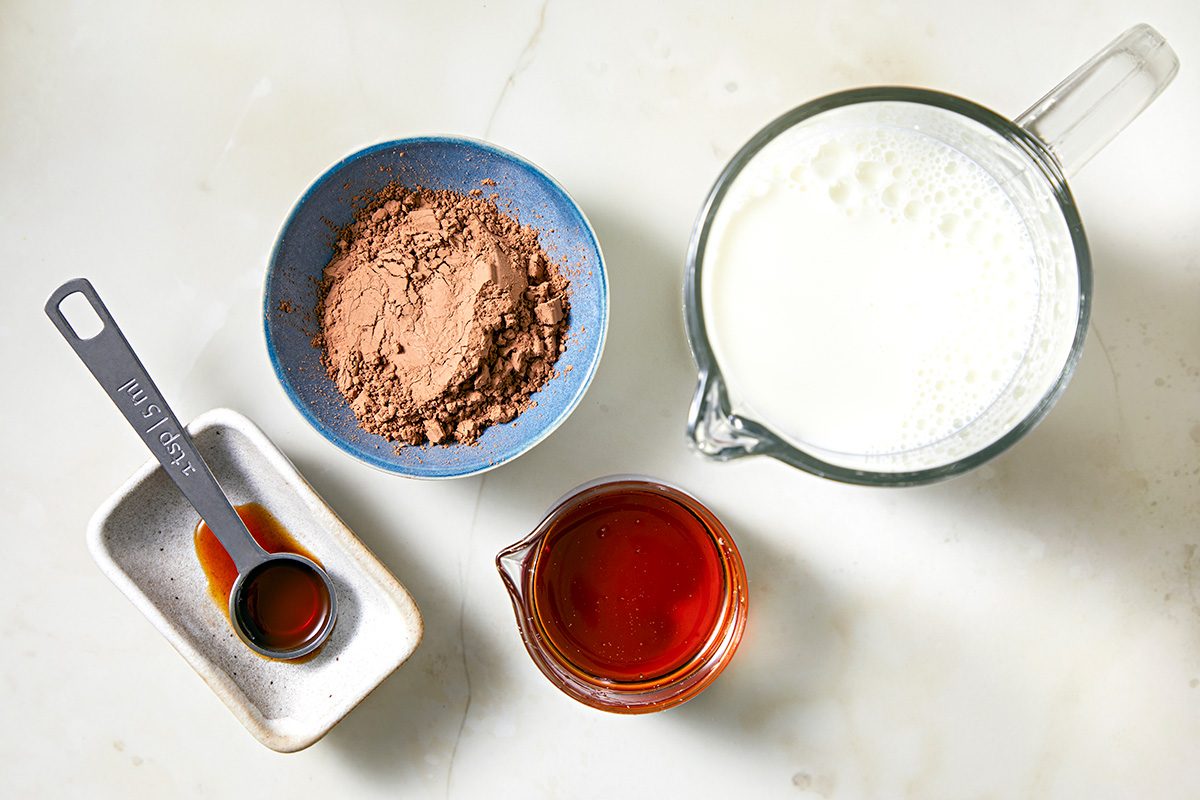 Ingredients for a recipe are displayed on a light surface: a blue bowl with cocoa powder, a glass measuring cup with milk, a small pitcher with reddish liquid, a rectangular dish with vanilla extract, and a metal measuring spoon.
