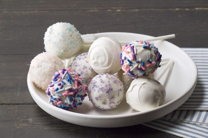 closeup shot of of cake pops; the pops are round and coated in various decorative toppings, they are presented on a simple white plate, in the background, a striped blue and white napkin