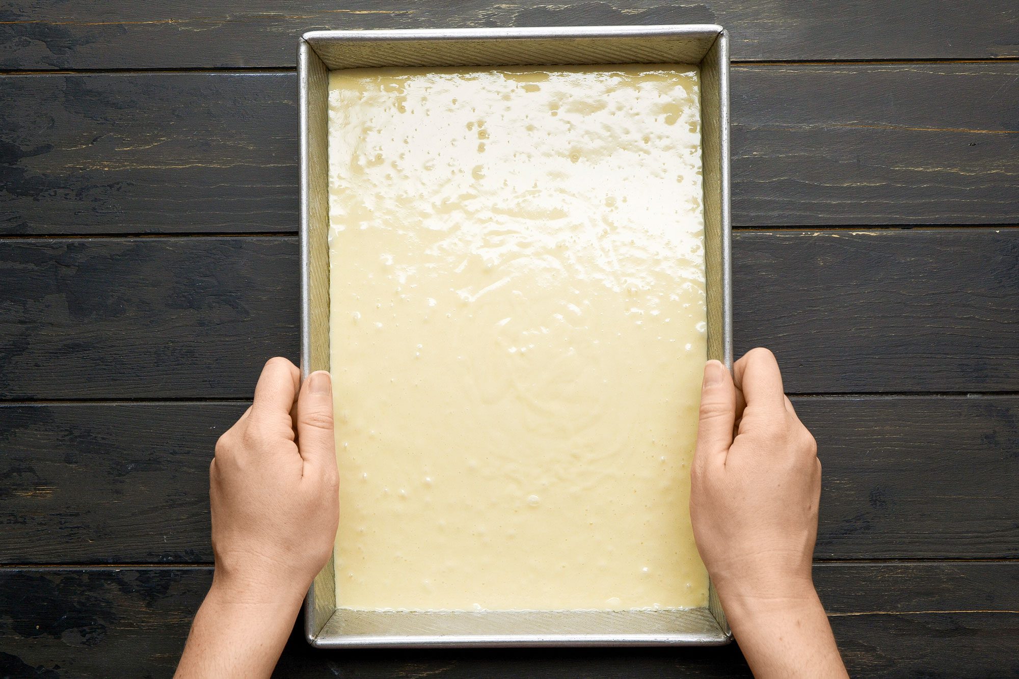 overhead shot of a rectangular baking pan filled with a creamy batter; Two hands are gently holding the pan from its sides; The pan is placed on a dark wooden surface