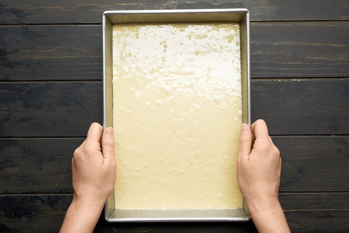 overhead shot of a rectangular baking pan filled with a creamy batter; Two hands are gently holding the pan from its sides; The pan is placed on a dark wooden surface