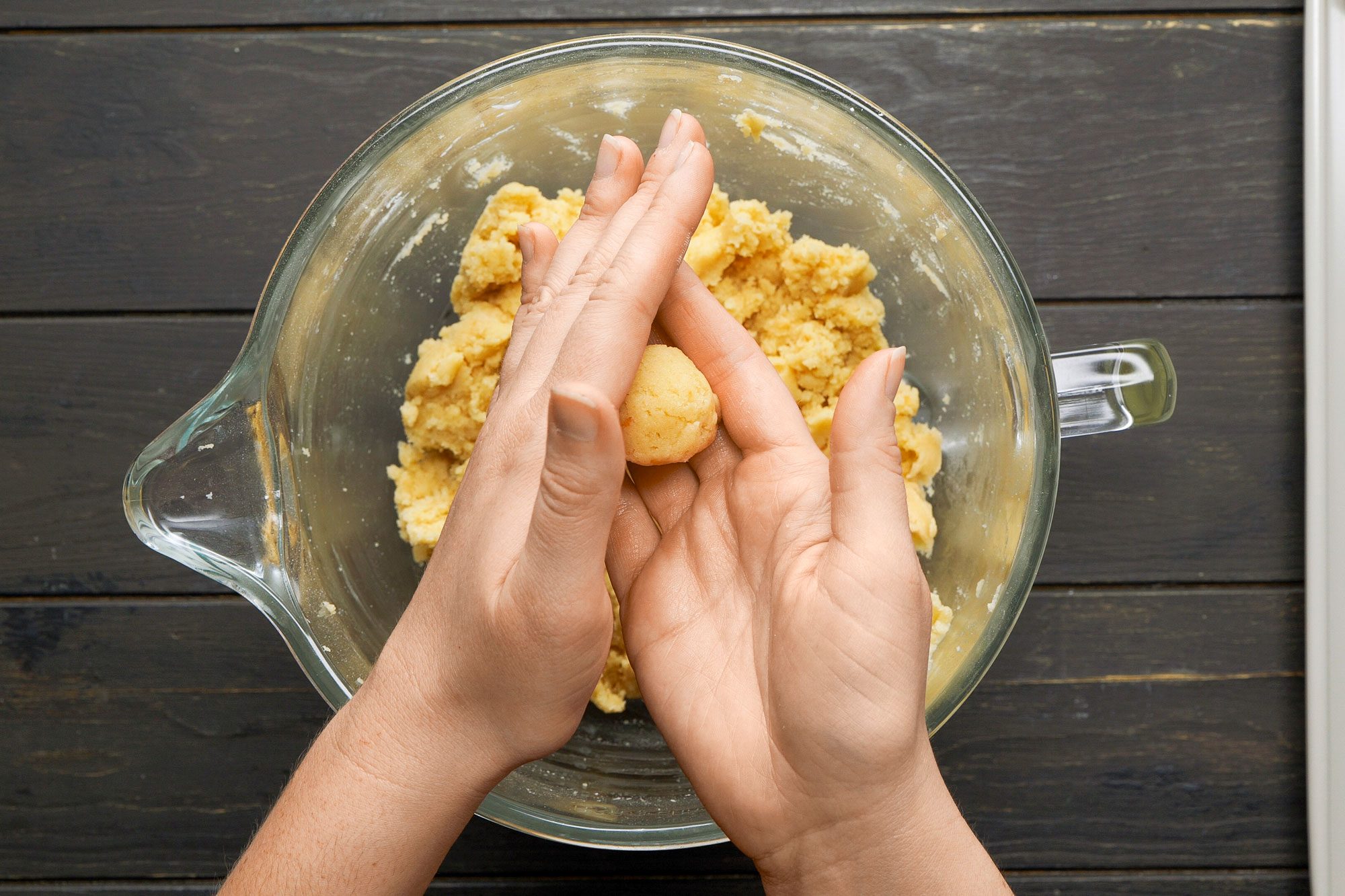 overhead shot of a pair of hands engaged in the process of rolling a small piece of dough in a glass mixing bowl, which contains a yellowish mixture in the background; the countertop beneath the bowl appears dark wood