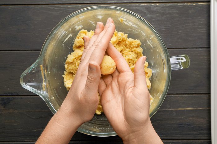 overhead shot of a pair of hands engaged in the process of rolling a small piece of dough in a glass mixing bowl, which contains a yellowish mixture in the background; the countertop beneath the bowl appears dark wood