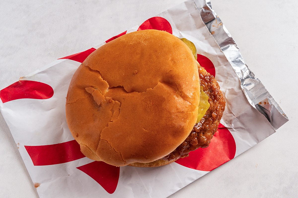 A chicken sandwich with a golden-brown bun, visible pickles, and a crispy chicken patty, placed on a foil-lined, branded wrapper.