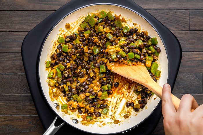 overhead shot of black beans being added to a white pan already containing sauteed green peppers, onions, and garlic