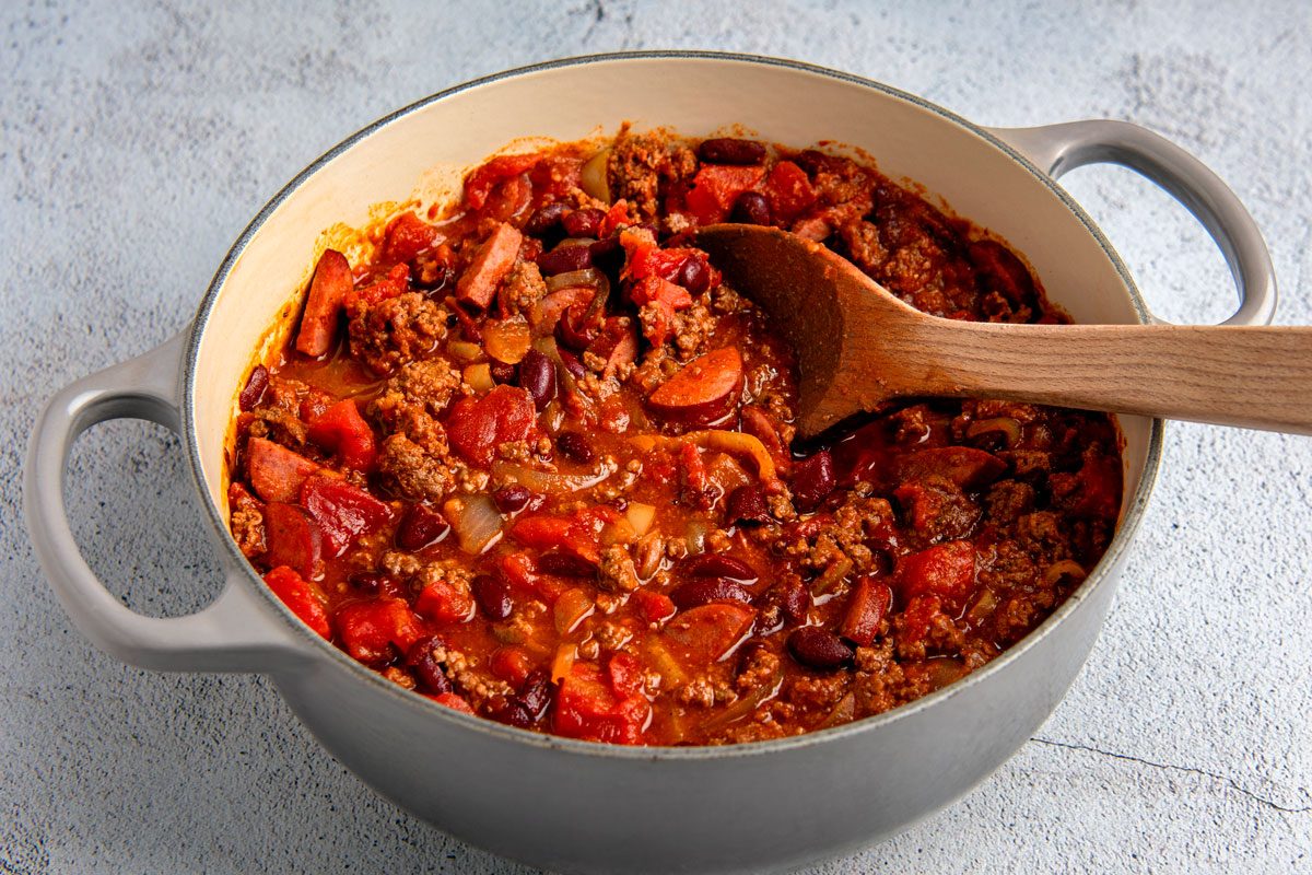 3/4th shot of a pot of chili with a wooden spoon in it, the pot is sitting on a light grey surface, the pot is grey with two handles