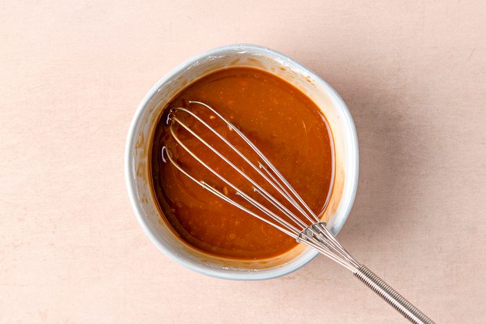 A small bowl filled with caramel sauce sits on a light surface. A metal whisk rests inside the bowl, partially submerged in the smooth, golden-brown caramel. The scene suggests preparation or cooking.