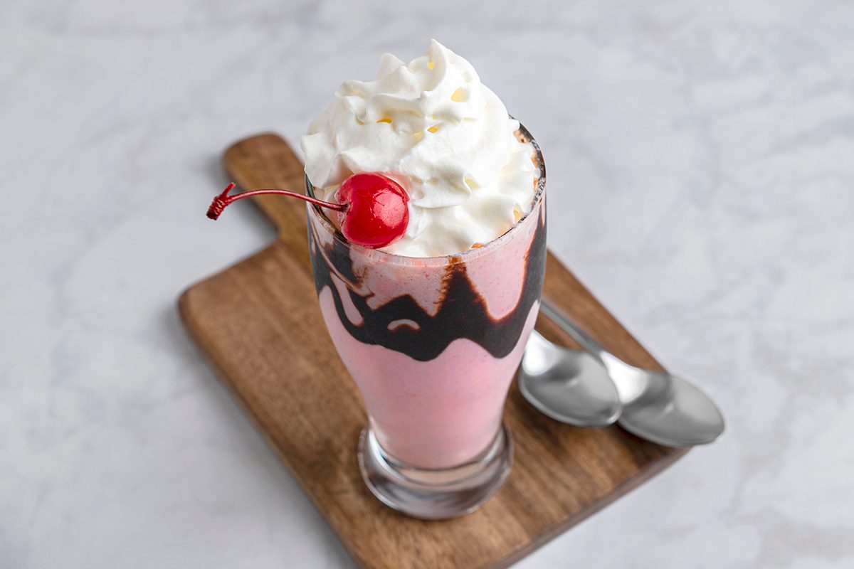 A tall glass of pink milkshake with whipped cream and a cherry on top, drizzled with chocolate syrup. It sits on a wooden board with two silver spoons beside it, all on a light gray marble surface.