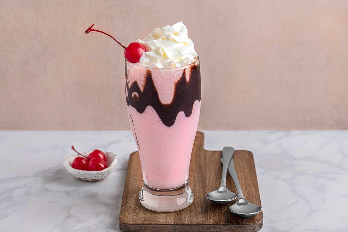 A tall glass of pink milkshake with chocolate drips and whipped cream, topped with a cherry, on a wooden board. Two silver spoons are beside it, and a small bowl of cherries is in the background on a marble surface.