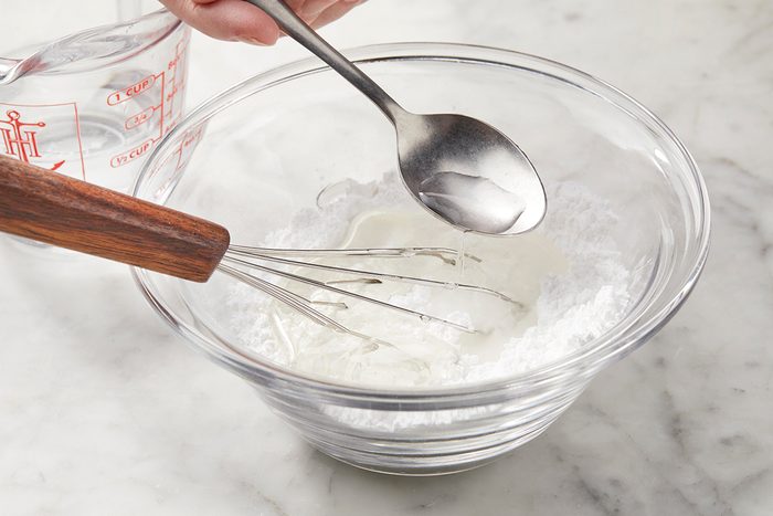 A glass bowl containing white powder with a whisk resting on its rim. A hand holds a spoon above the bowl, while another hand pours liquid from a measuring cup into the bowl. The scene is on a marble countertop.