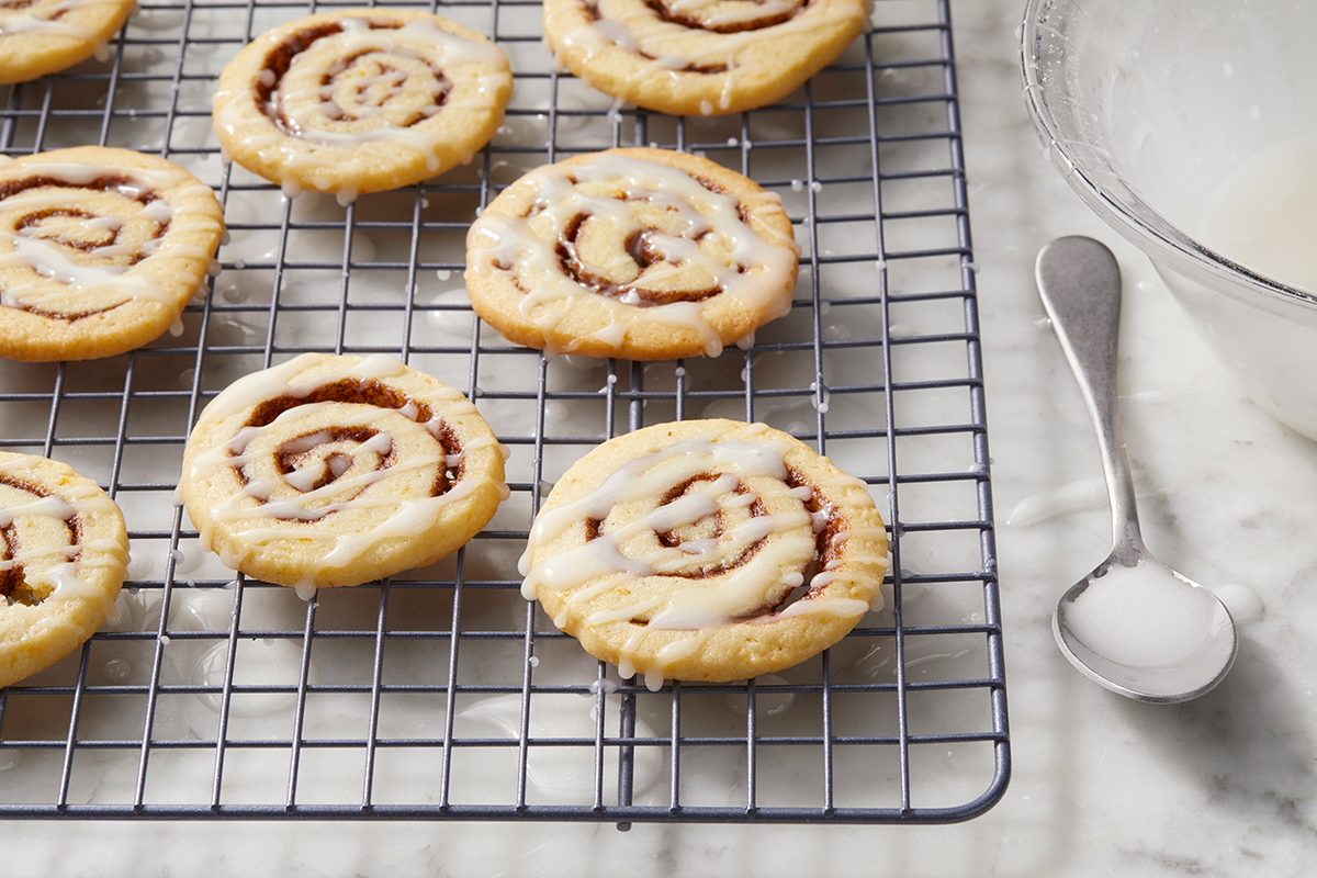 Cinnamon roll cookies with icing sit on a cooling rack. A glass bowl with a spoon beside it rests on a marble countertop.