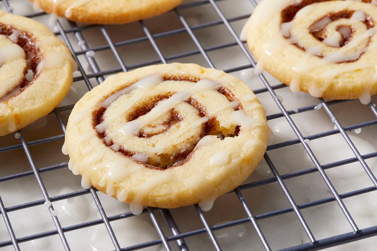 Close-up of cinnamon roll cookies with white icing drizzled on top, placed on a wire cooling rack. The cookies have a spiral design with a light brown and cream color, resting on a white surface.