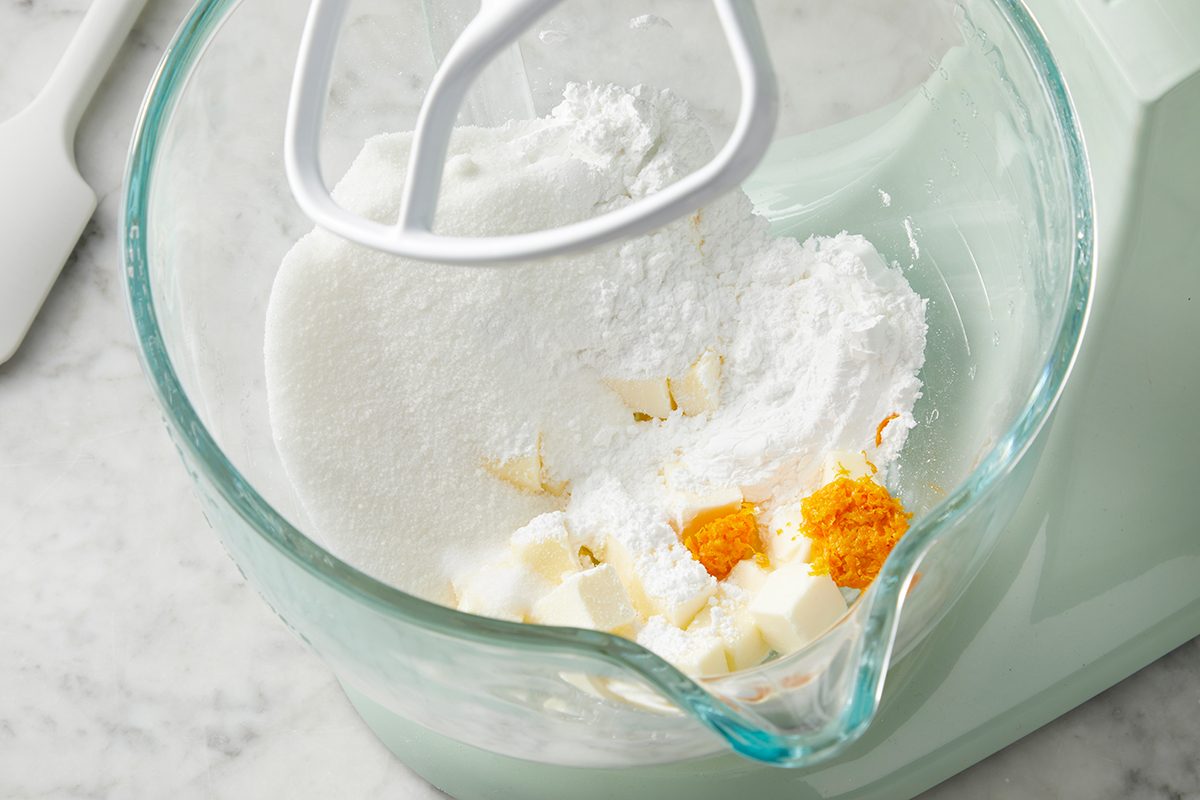 A mixing bowl with a paddle attachment contains powdered sugar, cubed butter, and small amounts of orange and yellow spices, set on a marble countertop. A spatula is visible in the background.