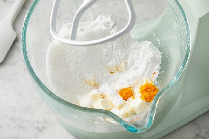 A mixing bowl with a paddle attachment contains powdered sugar, cubed butter, and small amounts of orange and yellow spices, set on a marble countertop. A spatula is visible in the background.