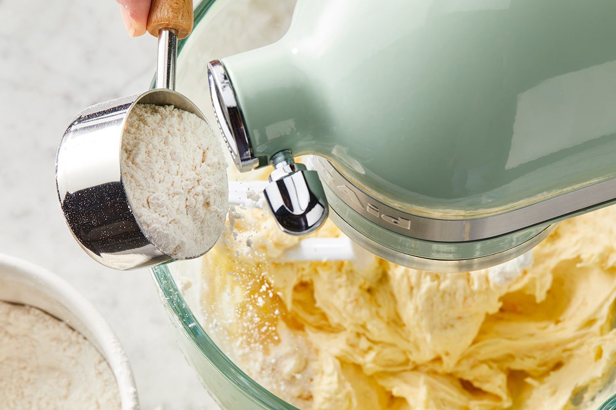 A person adds a cup of flour into a stand mixer with creamy batter. The mixer has a light green body, and the bowl is clear, showing the batter inside. A small white bowl with flour is next to the mixing bowl.