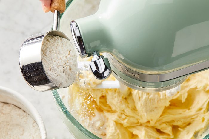A person adds a cup of flour into a stand mixer with creamy batter. The mixer has a light green body, and the bowl is clear, showing the batter inside. A small white bowl with flour is next to the mixing bowl.