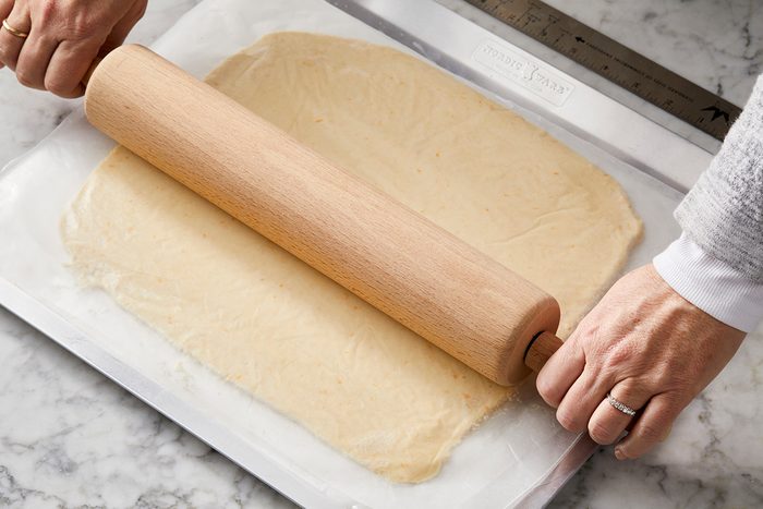 Hands rolling dough on a white surface with a wooden rolling pin. The dough is thin and spread out, and the rolling pin is being held by both hands.