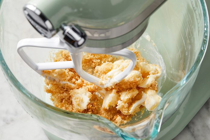 A stand mixer with a glass bowl is mixing butter and brown sugar. The ingredients are partially combined, indicating the beginning stages of a baking recipe. The mixer is set on a marble countertop.
