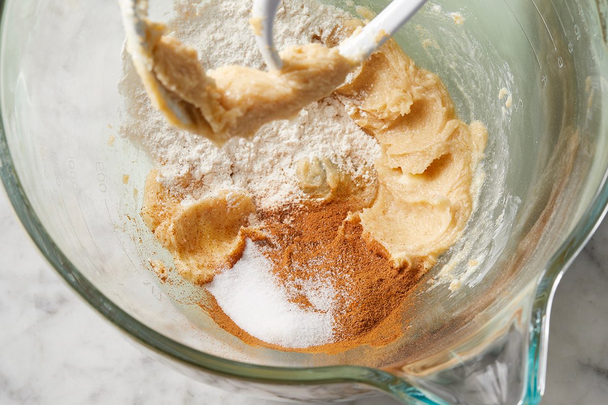Mixing bowl with cookie dough ingredients, including flour, sugar, cinnamon, and butter, partially mixed. A mixer attachment is visible, and the clear bowl rests on a marble countertop.