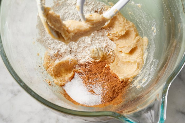 Mixing bowl with cookie dough ingredients, including flour, sugar, cinnamon, and butter, partially mixed. A mixer attachment is visible, and the clear bowl rests on a marble countertop.