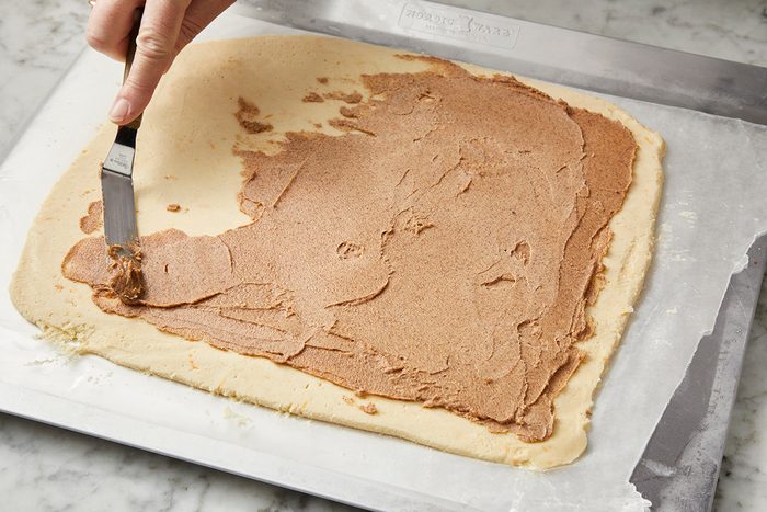 A hand spreads brown filling onto a rectangular sheet of dough with a spatula on a parchment-lined surface.