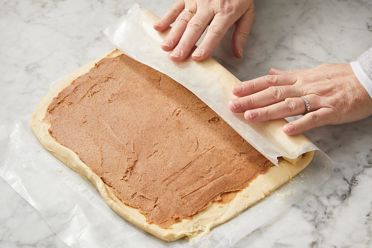 Hands are rolling up dough spread with a brown filling on a marble surface, using wax paper to assist in rolling. The dough is being prepared to make a rolled pastry or dessert.